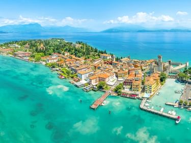 Luftaufnahme der Halbinsel Sirmione mit historischen Gebäuden und orangefarbenen Dächern, umgeben vom türkisfarbenen Wasser des Gardasees.