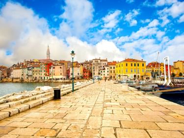 Steinerner Pier im Hafen von Poreč mit bunten historischen Gebäuden und Kirchturm. Boote rechts vertäut, blauer Himmel mit weißen Wolken.