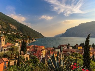 Dorf Torbole am Gardasee mit rotgedeckten Häusern, Kirchturm und Bergen. Sonnenuntergangslicht beleuchtet die Stadt am See und das Wasser.
