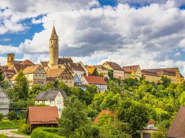 Blick auf die Stadt Kirchberg mit Fachwerkhäusern und hohem Kirchturm auf einem Hügel. Grüne Bäume im Vordergrund, dramatischer Wolkenhimmel.