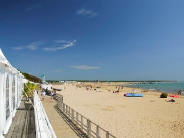Sandstrand von La Tranche-sur-Mer mit weißen Strandhütten entlang einer Holzpromenade. Strandbesucher entspannen auf dem Sand unter blauem Himmel.