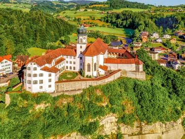 Luftaufnahme des Klosters Werthenstein mit weißen Gebäuden, roten Ziegeldächern und Kirchturm, umgeben von grünen Hügeln und Dorfhäusern.