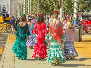 Vier Frauen in traditionellen Flamenco-Kleidern gehen von der Kamera weg. Kleider in Türkis, Rot und Grün mit Rüschen. Pferdekutschen im Hintergrund.