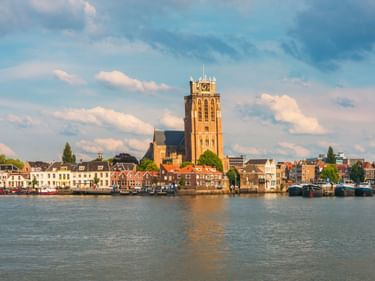 Blick auf die Uferpromenade von Dordrecht mit dem hohen Turm der Grote Kerk. Historische Gebäude säumen den Hafen mit Booten am Kai.