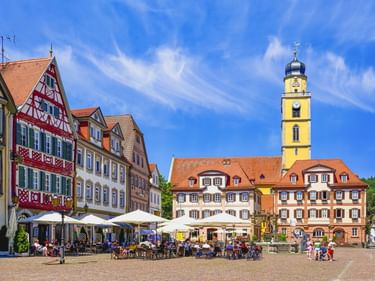 Historischer Marktplatz in Mergentheim mit bunten Fachwerkhäusern, Straßencafés, gelbem Kirchturm und Brunnen unter blauem Himmel.