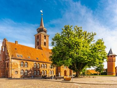 Rote Backsteinkirche mit orangefarbenem Ziegeldach und Turm in Nykøbing Falster. Großer Baum und Rundturm auf Kopfsteinpflasterplatz unter blauem Himmel.
