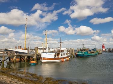 Hafen in Wyk auf Föhr mit festgemachten Booten, darunter ein weiß-braunes Schiff und türkisfarbenes Fischerboot. Rot-weißer Leuchtturm im Hintergrund unter blauem Himmel.
