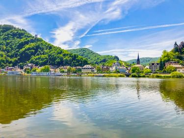 Blick auf Traben-Trarbach an der Mosel mit bewaldeten Hügeln, Kirchturm und historischen Gebäuden, die sich im ruhigen Wasser spiegeln.