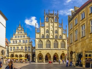 Gotisches Rathaus von Münster mit verzierter Fassade und Spitzbögen auf einem Kopfsteinpflasterplatz, umgeben von historischen Gebäuden.