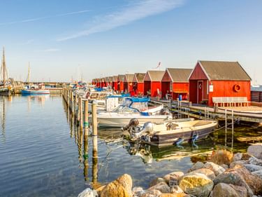 Hafen Rødvig mit roten Holz-Bootshäusern auf Holzstegen, kleine Boote im ruhigen Wasser, Steine im Vordergrund, Segelboote im Hintergrund.