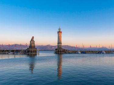 Lindauer Hafeneinfahrt mit Leuchtturm und bayerischem Löwen auf Steinsockeln im Bodensee, spiegelnd im ruhigen Wasser bei Sonnenuntergang.