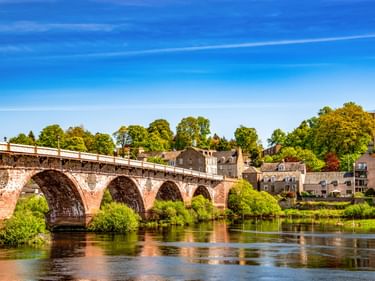 Historische Steinbrücke mit mehreren Bögen über den River Tay in Perth. Grüne Bäume und Wohngebäude am Flussufer unter blauem Himmel.