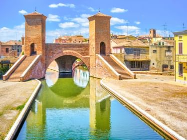 Historische Trepponti-Brücke in Comacchio mit zwei Ziegeltürmen und Treppen über einem grünen Kanal, umgeben von bunten italienischen Häusern.