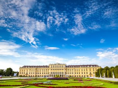 Gelbes Barockschloss mit Ziergärten mit geometrischen Blumenbeeten in Rot, Weiß und Grün unter blauem Himmel mit weißen Wolken.