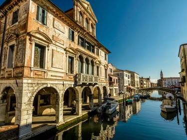 Kanal in Chioggia mit vertäuten Booten, die sich im ruhigen Wasser spiegeln. Historisches Gebäude mit Arkaden links, bunte Häuser säumen den Wasserweg.