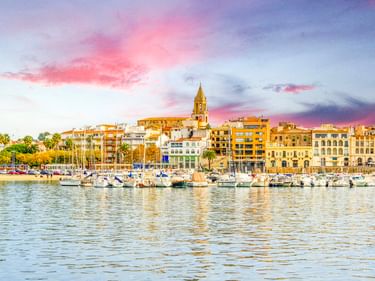 Yachthafen von Palamós mit weißen Booten im ruhigen Wasser. Bunte Gebäude und ein Kirchturm erheben sich hinter Palmen unter rosa-blauem Abendhimmel.