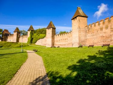 Historische Festungsmauern mit Türmen in Nymburk, Tschechien. Ein gepflasterter Weg führt durch grüne Wiese mit Bänken unter blauem Himmel.