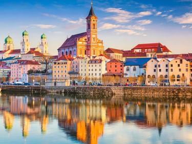Bunte historische Gebäude und Kirchtürme von Passau spiegeln sich bei Sonnenuntergang in der Donau unter blauem Himmel mit Wolken.