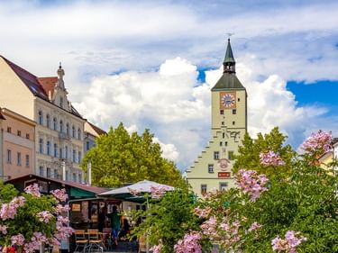Historischer Uhrturm in der Deggendorfer Altstadt mit bunten Gebäuden, rosa blühenden Sträuchern und Straßencafé unter blauem Himmel.