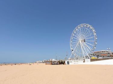 Weißes Riesenrad am Sandstrand von Saint-Jean-de-Monts unter klarem blauen Himmel, mit Strandgebäuden im Hintergrund.