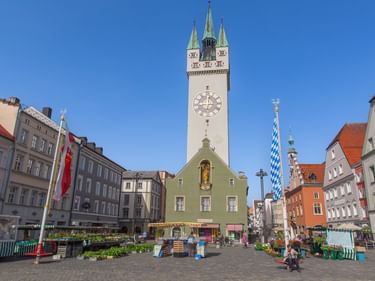 Straubinger Stadtplatz mit hohem Stadtturm, grünem Gebäude, bayerischer Flagge und bunten historischen Gebäuden unter blauem Himmel.