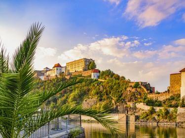 Veste Oberhaus auf einem Hügel über der Donau in Passau. Palmwedel rahmen die Ansicht mit historischen Gebäuden und blauem Himmel.