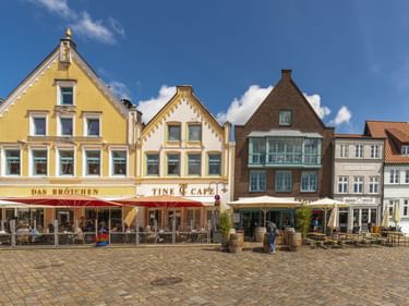 Bunte historische Gebäude mit Cafés und Außenbestuhlung auf einem Kopfsteinpflasterplatz in Husums Altstadt unter blauem Himmel.