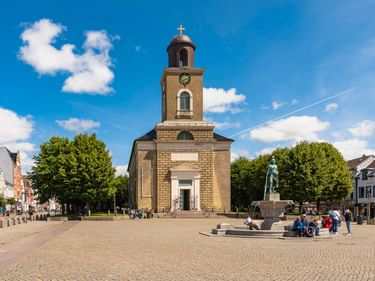 Husumer Marktplatz mit Steinkirche mit Glockenturm, zentralem Brunnen mit Statue, Kopfsteinpflaster und umliegenden Gebäuden unter blauem Himmel.