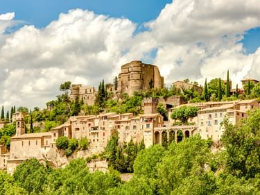 Montbrun-les-Bains, ein mittelalterliches Bergdorf mit Steinhäusern und Burgruine, umgeben von Zypressen und üppigem Grün unter blauem Himmel.