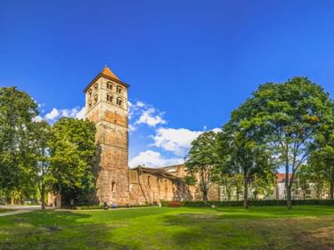 Steinturm und Ruinen der Stiftsruine Bad Hersfeld, umgeben von grüner Wiese und Bäumen unter blauem Himmel. Historische romanische Kirchenreste.