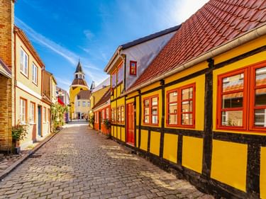 Kopfsteinpflasterstraße in Faaborg mit bunten Fachwerkhäusern. Ein gelbes Haus mit roten Fenstern ist prominent, im Hintergrund ist ein Kirchturm sichtbar.