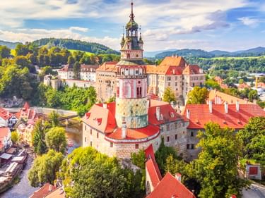 Luftaufnahme von Český Krumlov mit Schlossturm, roten Ziegeldächern und grünen Hügeln unter blauem Himmel.