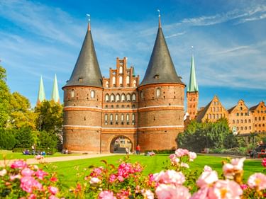 Historisches Holstentor in Lübeck mit zwei Kegeltürmen und roter Backsteinfassade. Rosa Blumen blühen im Vordergrund, grüner Rasen und Kirchtürme sichtbar.