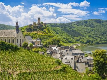 Beilstein mit Kirche und Burg Metternich auf dem Hügel, Weinberge im Vordergrund, Mosel und grüne Hügel unter blauem Himmel.