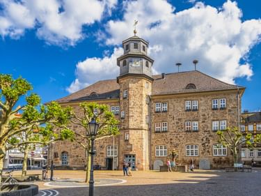 Steinernes Rathausgebäude in Witzenhausen mit Uhrturm und weißer Kuppel, umgeben von Bäumen auf einem sonnigen Platz mit blauem Himmel.