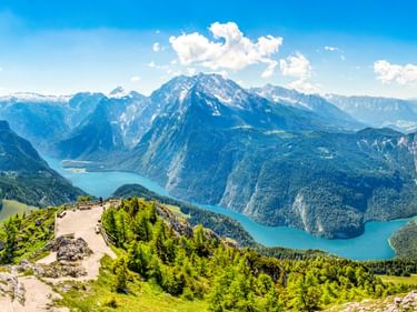 Luftaufnahme des türkisfarbenen Königssees, umgeben von steilen Bergen. Grüne Wiesen und Wälder im Vordergrund, schneebedeckte Gipfel.