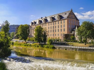 Welfenschloss, ein großes Renaissance-Schloss mit Gauben in Hann. Münden, vom Fluss mit Wehr aus gesehen. Bäume säumen das Flussufer.