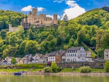 Schloss Stolzenfels thront auf einem bewaldeten Hang über der Mosel bei Koblenz, mit einem Flussdorf darunter und blauem Himmel darüber.