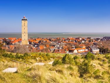 Brandaris Leuchtturm überragt das Dorf Terschelling mit orangefarbenen Ziegeldächern, gesehen von Dünengras. Blauer Himmel und Wasser im Hintergrund.