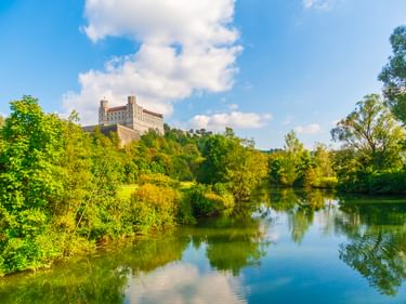 Willibaldsburg auf einem Hügel in Eichstätt mit Blick auf einen ruhigen Fluss, umgeben von grünen Bäumen unter blauem Himmel mit weißen Wolken.