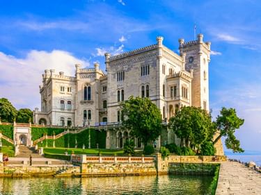Weißes Schloss Miramare in Triest mit terrassierten Gärten zur Adria unter blauem Himmel mit weißen Wolken.