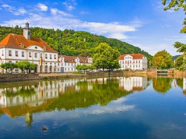 Weiße Barockgebäude mit orangen Dächern spiegeln sich in der ruhigen Weser in Bad Karlshafen, umgeben von grünen bewaldeten Hügeln.