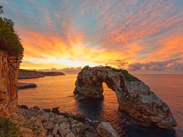 Natürlicher Felsbogen im Mittelmeer bei Sonnenuntergang auf Mallorca, mit dramatischen orange-rosa Wolken, die sich im Wasser spiegeln.