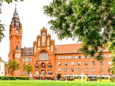 Rotes Backsteinrathaus Köpenick mit gotischem Turm und Uhr, eingerahmt von grünen Bäumen. Gelbe Straßenbahnen fahren vorbei.