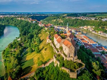Luftaufnahme der Burghausen Altstadt mit mittelalterlicher Burg auf Hügel, orangefarbenen Dächern, Fluss und grünen Wäldern.