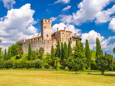 Mittelalterliches Schloss Fagagna mit Steinmauern und Turm auf grünem Hügel, umgeben von Zypressen und Rasen unter blauem Himmel mit weißen Wolken.