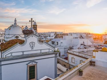 Weiße Kirche mit Kuppel und Kreuz überblickt weiß getünchte Dächer in Olhão bei Sonnenuntergang. Orangefarbener Himmel und traditionelle portugiesische Architektur sichtbar.