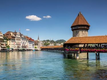 Historische Holz-Kapellbrücke mit achteckigem Wasserturm über der Reuss in Luzern, mit Blumen geschmückt, Altstadthäuser sichtbar.