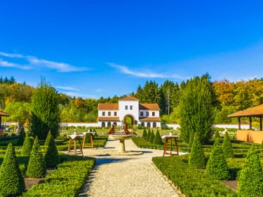 Weiße Villa im römischen Stil mit rotem Ziegeldach in Villa Borg, umgeben von gepflegten Hecken und formalen Gärten mit Kiesweg zum Eingang.