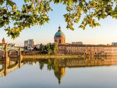 Blick auf Toulouse mit der Garonne, die die rosa Kuppel des Hôpital de La Grave spiegelt. Saint-Pierre-Brücke links, eingerahmt von grünen Blättern.